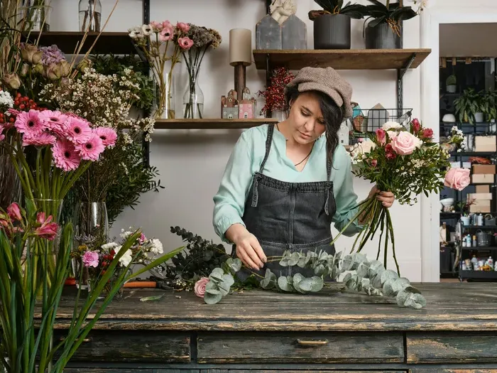 Woman florist arranging a bouquet of pink roses and greenery behind a rustic counter in a flower shop